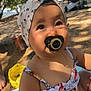 baby, pacifier, headband, child, outdoor, sunlight, tree, stroller, necklace, summer_clothing, floral_pattern, skin, face, eyes, cute, infant, nature, ground, shade, portrait
