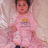 baby, child, infant, portrait, sitting, curly_hair, pink_clothing, onesie, smiling, serious_expression, pillow, blanket, bed, balloon, sock, indoors, bedroom, furniture, photograph, text_on_clothing