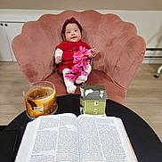 Ailany is registered to the contest to win money with this photo: baby, child, red_clothing, headband, pink_flowers, pink_chair, plush_chair, open_book, candle, sunflower_candle, miniature_house, table, wooden_floor, indoor, smiling, sitting, decor, cozy, warm_lighting, cute