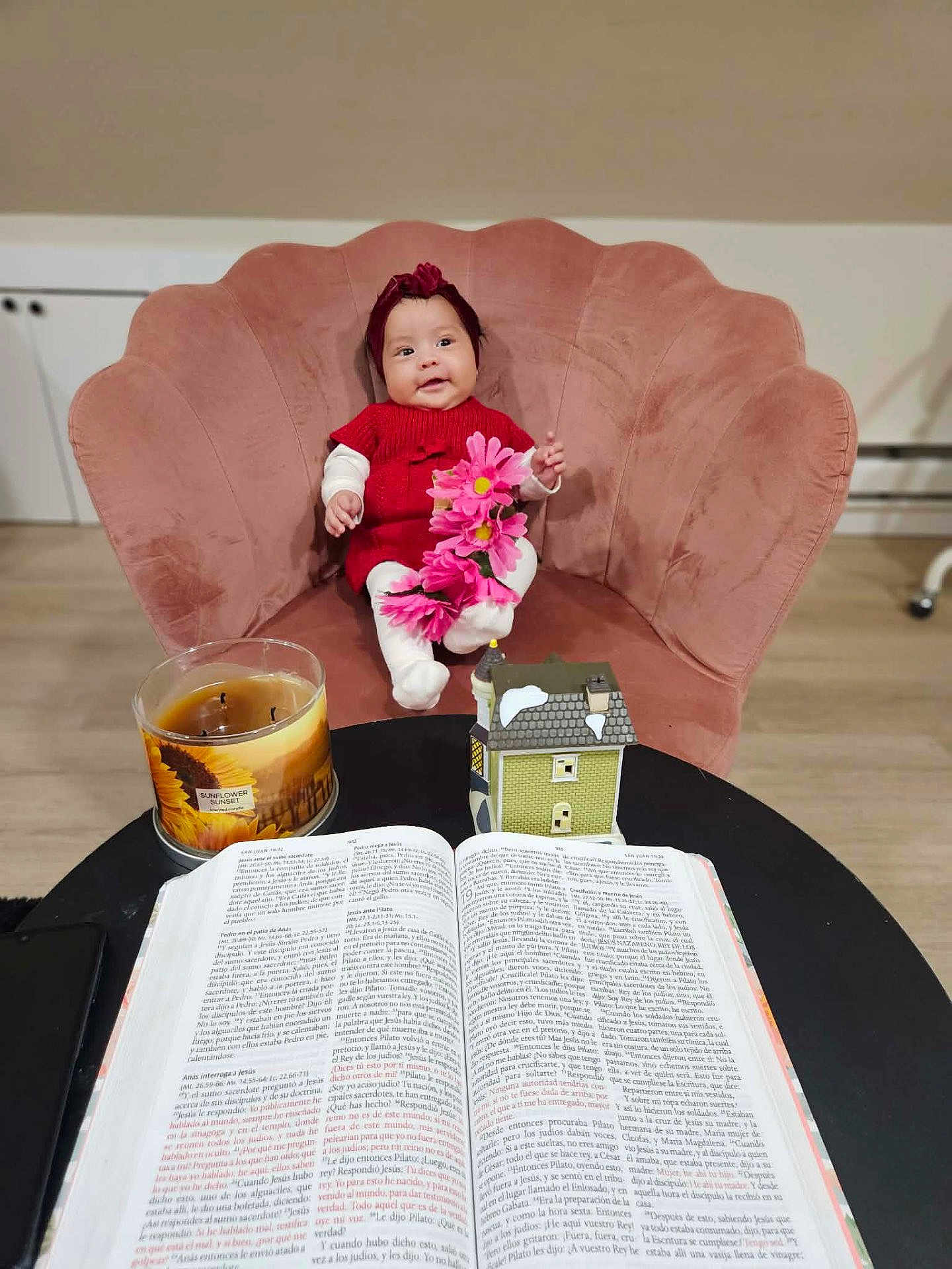 Ailany is registered to the contest to win money with this photo: baby, child, red_clothing, headband, pink_flowers, pink_chair, plush_chair, open_book, candle, sunflower_candle, miniature_house, table, wooden_floor, indoor, smiling, sitting, decor, cozy, warm_lighting, cute