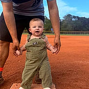 Lawson is registered to the contest to win money with this photo: baby, child, infant, overalls, socks, adult_hands, baseball_field, baseball_base, outdoor, grass, sky, green_overalls, smiling, standing, person, helping_hand, seven_months, sign, sunlight, daytime