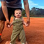 baby, child, infant, overalls, socks, adult_hands, baseball_field, baseball_base, outdoor, grass, sky, green_overalls, smiling, standing, person, helping_hand, seven_months, sign, sunlight, daytime
