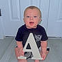 Lawson is registered to the contest to win money with this photo: baby, child, infant, letter_a, black_clothing, blue_eyes, sitting, barefoot, wooden_floor, door, indoors, smiling, cute, holding_object, young_child, portrait, light_skin, short_hair, playful, front_view