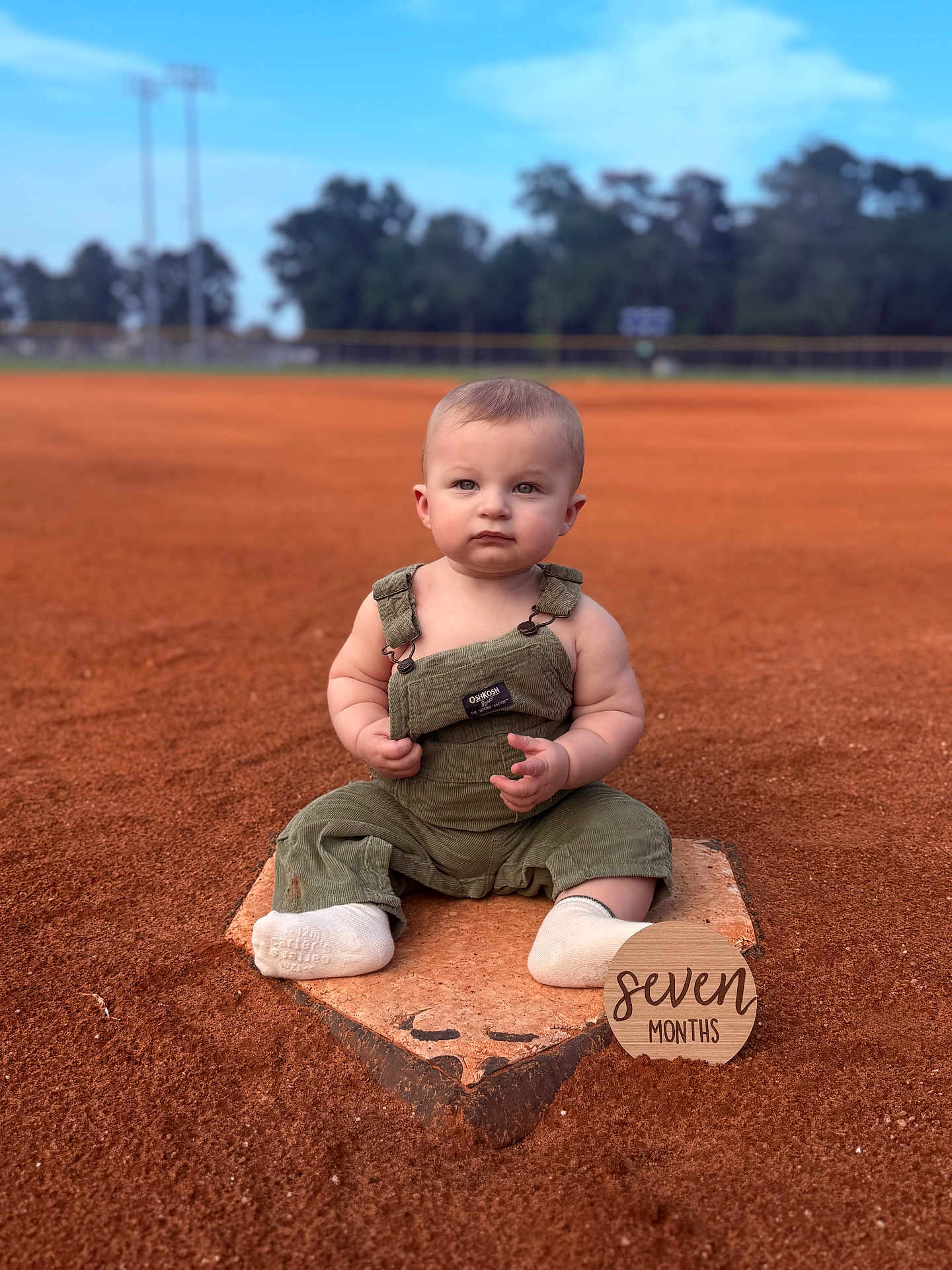 Lawson is registered to the contest to win money with this photo: baby, child, overalls, baseball_field, base, dirt, outdoor, milestone, sign, seven_months, green_clothing, socks, infant, grass, trees, sky, portrait, sitting, cute, daylight