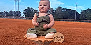 Lawson is registered to the contest to win money with this photo: baby, infant, child, overalls, baseball_field, home_plate, outdoor, sky, trees, grass, dirt, sign, milestone, socks, cute, portrait, smiling, fist, daylight, nature