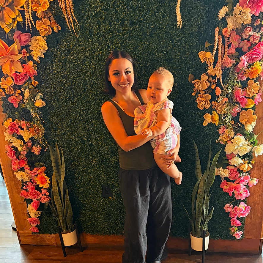 Celia is registered to the contest to win money with this photo: woman, baby, child, flower_wall, plants, greenery, smile, indoor, portrait, casual_clothing, happy, standing, decor, pot_plants, wood_floor, colorful, nature_inspired, mother_and_child, family, people