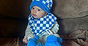 Zander is registered to the contest to win money with this photo: baby, child, blue_hat, blue_booties, checkered_pattern, bib, couch, brown_couch, indoor, cute, infant, sitting, clothing, footwear, portrait, young_child, cozy, soft_toys, home, curious