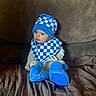 baby, child, blue_hat, blue_booties, checkered_pattern, bib, couch, brown_couch, indoor, cute, infant, sitting, clothing, footwear, portrait, young_child, cozy, soft_toys, home, curious