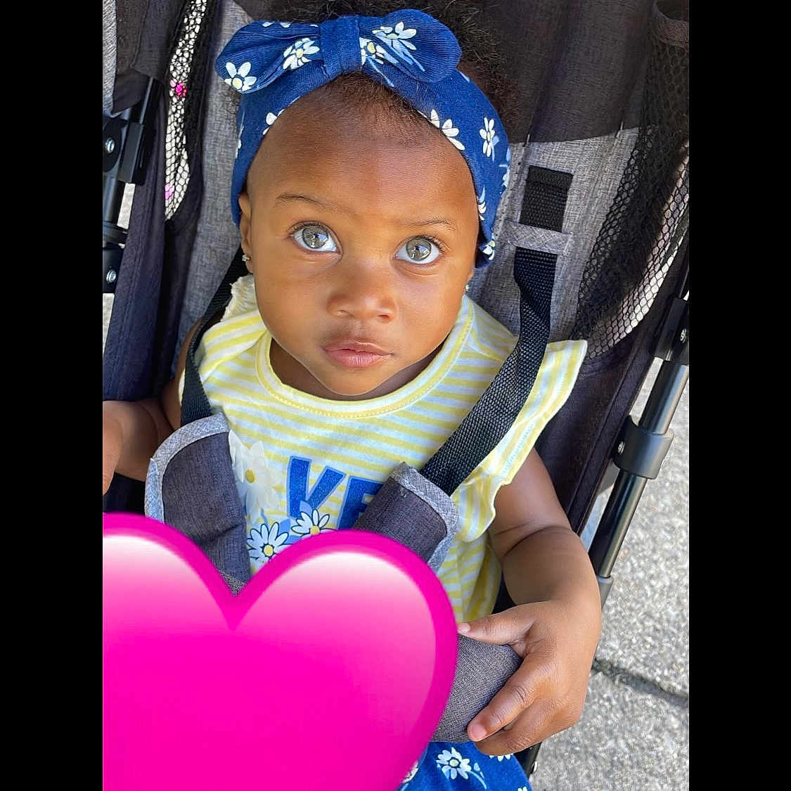 Jewel is registered to the contest to win money with this photo: baby, blue_eyes, blue_skirt, child, curious, cute, expression, face, floral_pattern, hand, headband, outdoor, pink_heart, portrait, seatbelt, skin, stroller, summer_clothing, toddler, yellow_shirt