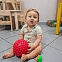 child, toddler, toy, red_ball, green_orange_toy, floor, tile_floor, wooden_chair, wooden_table, basket, vegetables, indoor, shirt, shorts, sitting, curious, play, furniture, person, baby