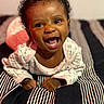 Thalia joined the competition — help win amazing prizes! baby, child, infant, smiling, laughing, portrait, curly_hair, dark_skin, eyes, open_mouth, onesie, floral_clothing, sitting, blanket, striped_blanket, bed, bedroom, indoor, happy, close_up