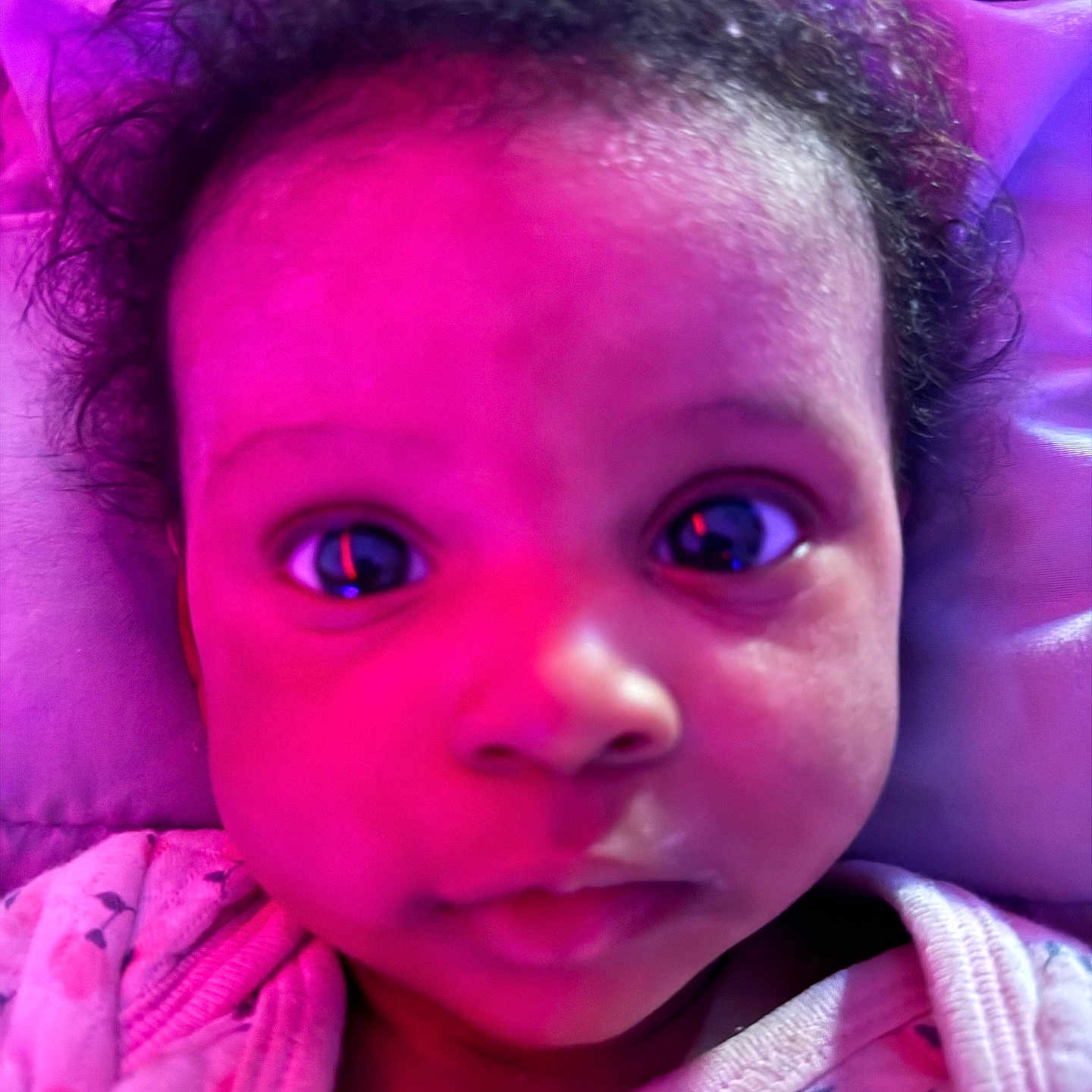 Thalia is registered to the contest to win money with this photo: baby, infant, child, portrait, close_up, face, wide_eyes, curly_hair, looking_at_camera, indoor, neon_lighting, purple_lighting, pink_lighting, blanket, clothing, patterned_fabric, cozy, headshot, bed, photography