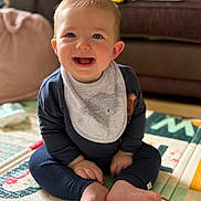 Ren is registered to the contest to win money with this photo: baby, child, smiling, sitting, play_mat, bib, clothing, foot, hand, living_room, couch, cushion, floor, indoors, happy, person, face, blue_eyes, hair, infant