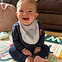 baby, child, smiling, sitting, play_mat, bib, clothing, foot, hand, living_room, couch, cushion, floor, indoors, happy, person, face, blue_eyes, hair, infant