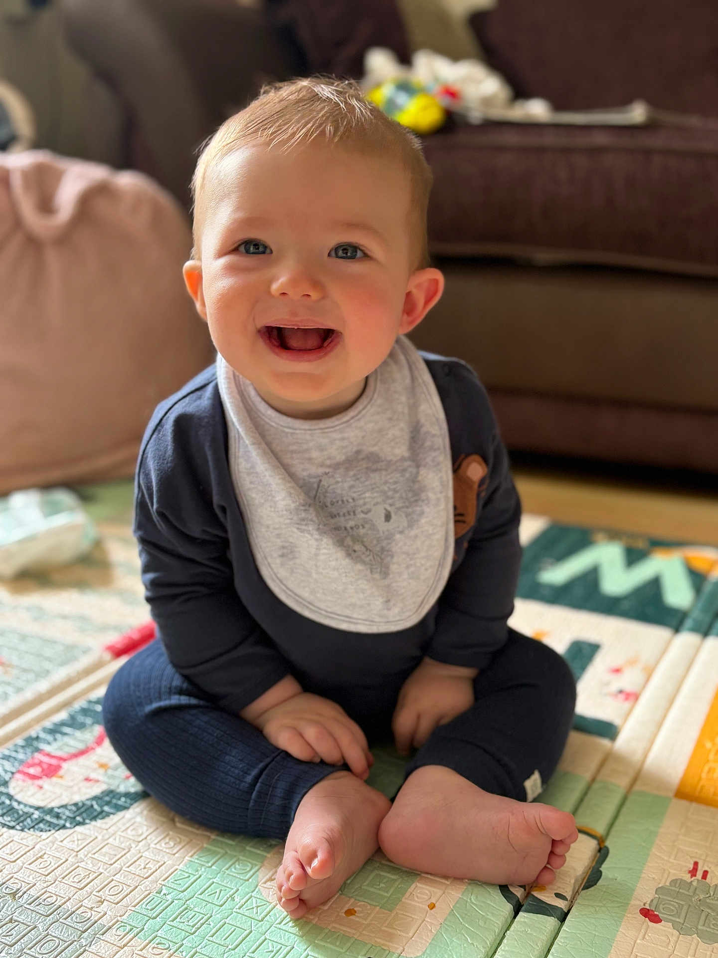 Ren is registered to the contest to win money with this photo: baby, child, smiling, sitting, play_mat, bib, clothing, foot, hand, living_room, couch, cushion, floor, indoors, happy, person, face, blue_eyes, hair, infant