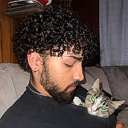 Shayla is registered to the contest to win money with this photo: man, curly_hair, beard, earring, cat, tabby_cat, indoor, couch, gray_couch, black_shirt, chain_necklace, curtain, wooden_wall, pet, holding, close_up, flash_reflection, relaxed, comfort, companion