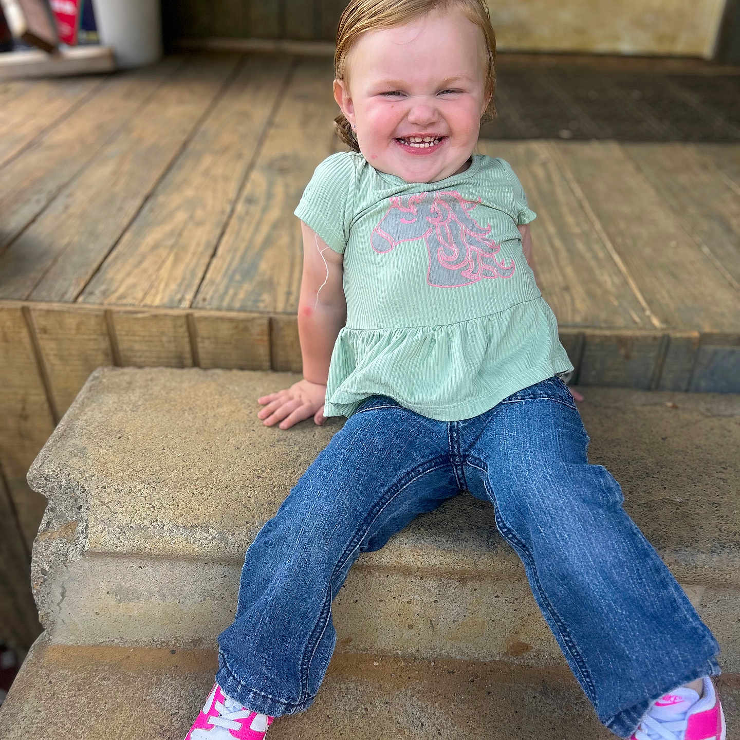 Katie Jo is registered to the contest to win money with this photo: casual_clothing, child, concrete_step, cute, daylight, green_shirt, happy, jeans, outdoor, person, pink_shoes, playful, portrait, sitting, smiling, sneakers, toddler, unicorn_print, wooden_floor, young_child