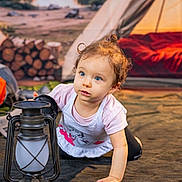 Viviana is registered to the contest to win money with this photo: toddler, child, lantern, camping, tent, firewood, outdoor, curly_hair, blue_eyes, crawling, adventure, nature, explorer, casual_clothing, soft_lighting, ground, playful, daylight, portrait, cute