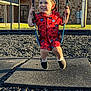 child, toddler, swing, playground, smile, cap, pajamas, red_clothing, shoes, socks, sunlight, shadow, rubber_chips, mat, grass, brick_wall, metal_structure, outdoor, candid, happy