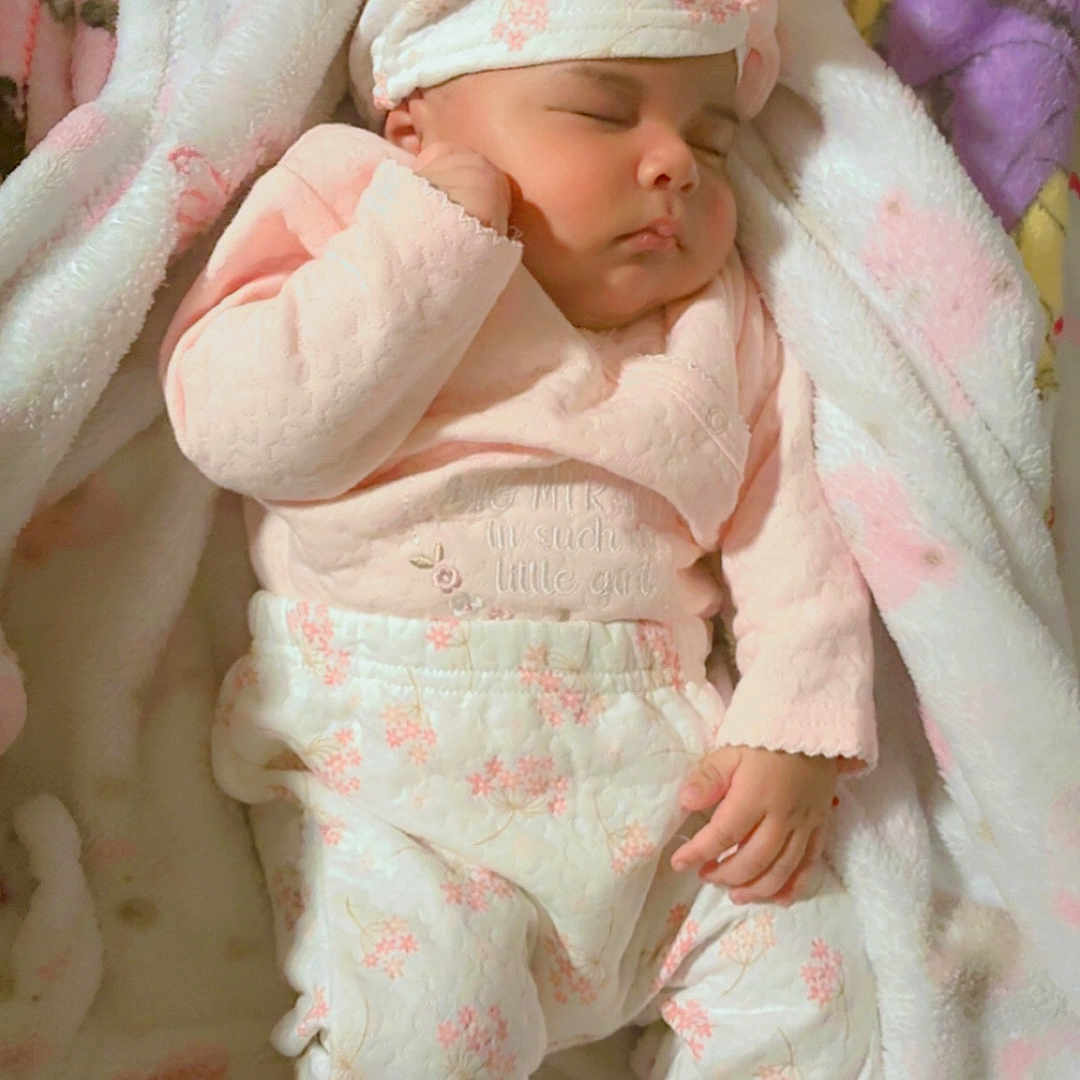 Sandra Gonzalez joined the competition — help win amazing prizes! baby, blanket, child, closeup, cozy, cute, floral_pattern, footed_pajamas, hat, headwear, indoor, infant, newborn, peaceful, person, pink_clothing, resting, sleeping, soft_texture, white_clothing