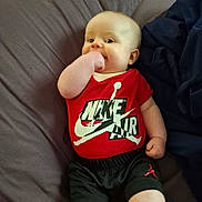 Dustin joined the competition — help win amazing prizes! baby, infant, child, smiling, lying_down, bed, blanket, red_shirt, nike_logo, brand, onesie, shorts, hand_in_mouth, cute, portrait, indoor, fabric, pillow, skin, eyes