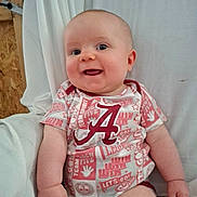 Dustin joined the competition — help win amazing prizes! baby, infant, smiling, onesie, logo, portrait, face, eyes, cheeks, arm, hand, happy, sitting, blanket, bed, curtain, wood_panel, bedding, cute, clothing_pattern