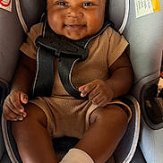 Marquiez is registered to the contest to win money with this photo: baby, child, car_seat, smiling, curly_hair, brown_outfit, white_socks, infant, seatbelt, happy, cute, indoors, portrait, young_child, sitting, person, skin, face, hands, feet