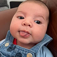 Mylo is registered to the contest to win money with this photo: baby, face, tongue, denim, overalls, mickey_mouse, clothing, child, cute, portrait, infant, closeup, expression, person, sitting, indoor, soft_light, skin, head, smile