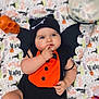 adorable, baby, bat_wings, black_hat, blanket, child, chubby_cheeks, costume, cute, decorations, festive, halloween, holiday, indoor, infant, orange_outfit, orange_skull, patterned_background, portrait, sucking_finger