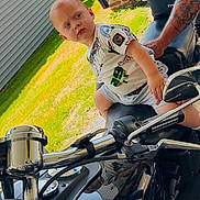 Ronan is registered to the contest to win money with this photo: toddler, motorcycle, man, tattoo, outdoor, grass, brick_pillar, garage, reflection, handlebars, shirt, shorts, sunlight, side_profile, person, vehicle, metal, expression, casual, summer