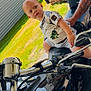 toddler, motorcycle, man, tattoo, outdoor, grass, brick_pillar, garage, reflection, handlebars, shirt, shorts, sunlight, side_profile, person, vehicle, metal, expression, casual, summer