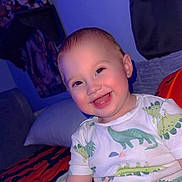 Ronan is registered to the contest to win money with this photo: baby, bedroom, blanket, child, clothing, cozy, cute, dinosaur_print, dressed, face, happy, indoor, infant, person, pillow, plush, seated, smiling, toy, young_child