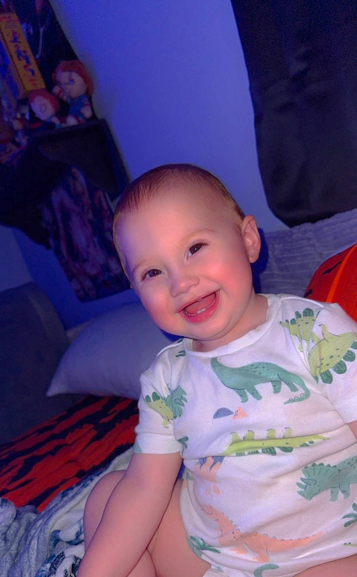 Ronan is registered to the contest to win money with this photo: baby, child, smiling, dinosaur_print, clothing, indoor, bedroom, blanket, pillow, toy, plush, happy, face, cute, infant, person, seated, young_child, dressed, cozy