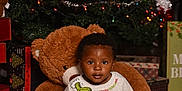 Omari is registered to the contest to win money with this photo: toddler, child, plush_toy, teddy_bear, christmas_tree, holiday_decor, presents, sweater, grinch, red_socks, pants, festive, indoors, smiling, fur_rug, lights, ornaments, ribbon, celebration, portrait