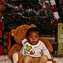 toddler, child, plush_toy, teddy_bear, christmas_tree, holiday_decor, presents, sweater, grinch, red_socks, pants, festive, indoors, smiling, fur_rug, lights, ornaments, ribbon, celebration, portrait
