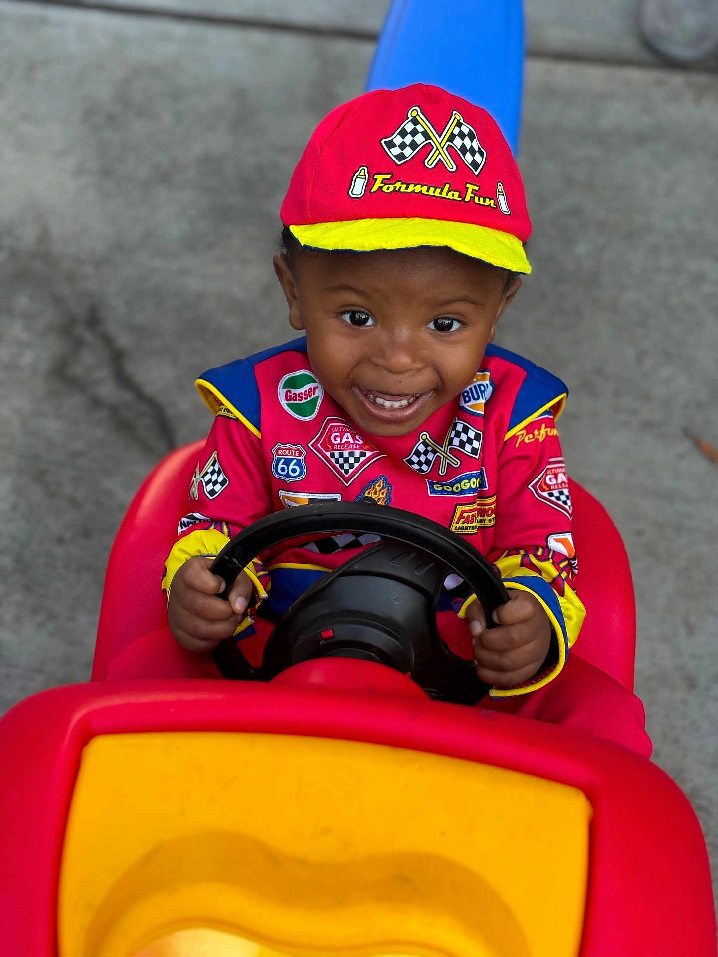 Omari is registered to the contest to win money with this photo: toddler, child, toy_car, red_clothing, cap, smiling, outdoor, steering_wheel, play, happy, race_car_driver, costume, young_child, hands, face, vehicle, concrete, bright_colors, hat, excitement