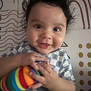 Valentino is registered to the contest to win money with this photo: baby, child, smiling, toy, rainbow, colorful, hands, curly_hair, face, cheeky, indoor, patterned_wall, geometric, clothing, portrait, cute, happy, young_child, playful, person
