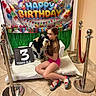 child, dog, birthday, indoor, balloons, sign, bench, floor, celebration, gift, banner, red_rope, stanchion, tile_floor, couch, smiling, hugging, colorful, festive, casual_clothing