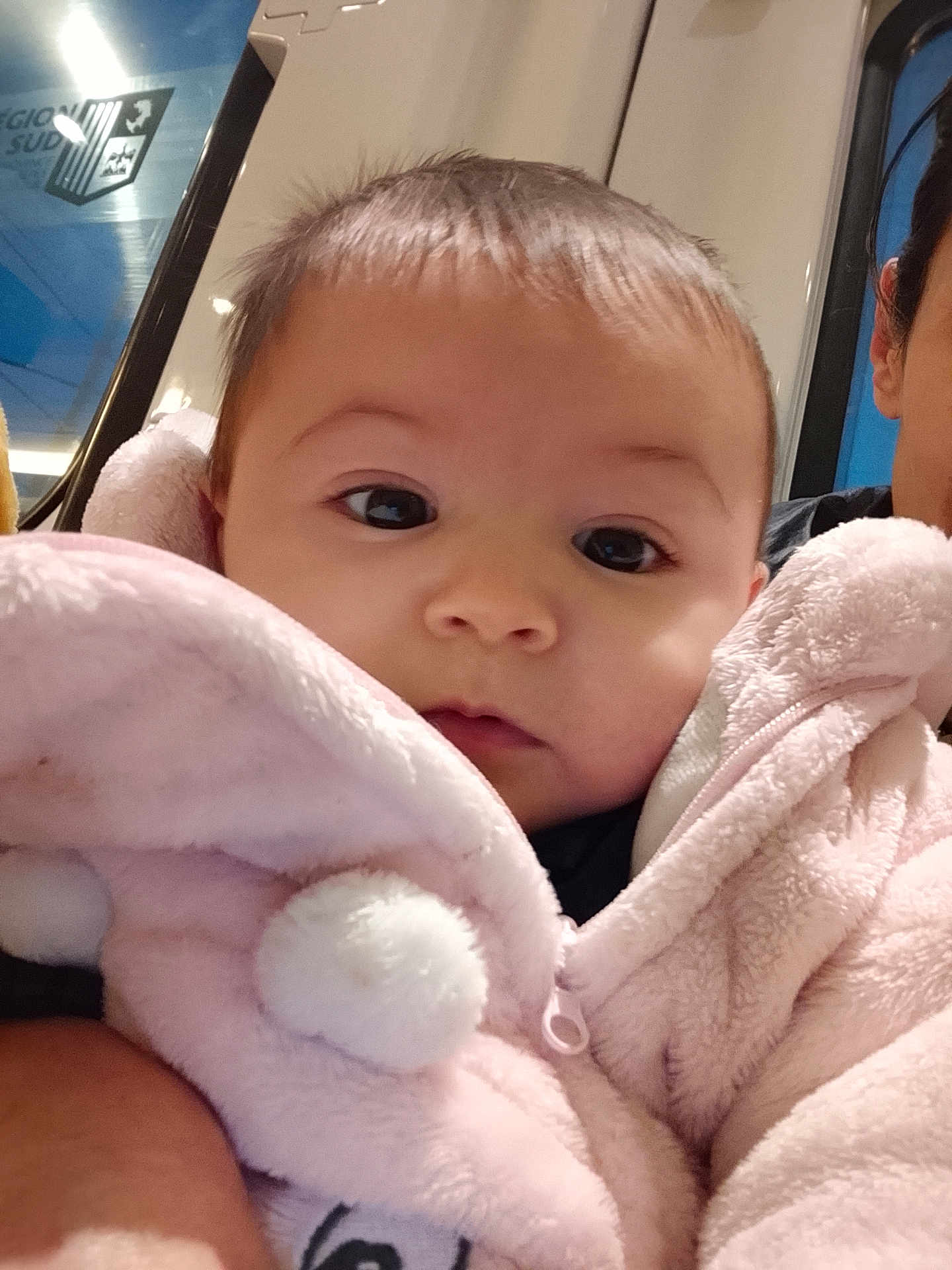 Leya a rejoint le concours — aidez-le/la à gagner de superbes lots ! baby, infant, child, portrait, close_up, pink_jacket, fluffy, pompom, face, eyes, cheeks, cozy, candid, parent_partial, public_transport, window, signage, soft_toy, hooded_jacket, indoor