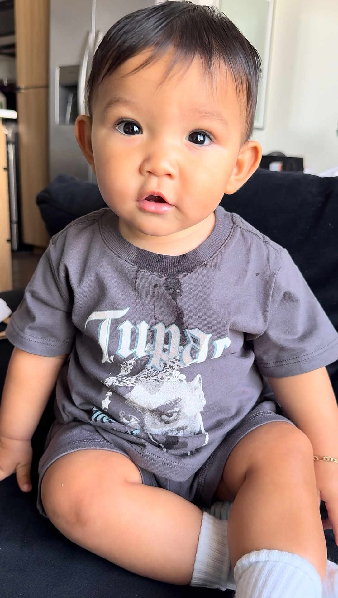 Santiago is registered to the contest to win money with this photo: toddler, child, indoor, portrait, sitting, tshirt, graphic_tee, spill, curious, face, eyes, shorts, socks, couch, furniture, home, young_child, expression, casual_clothing, bracelet