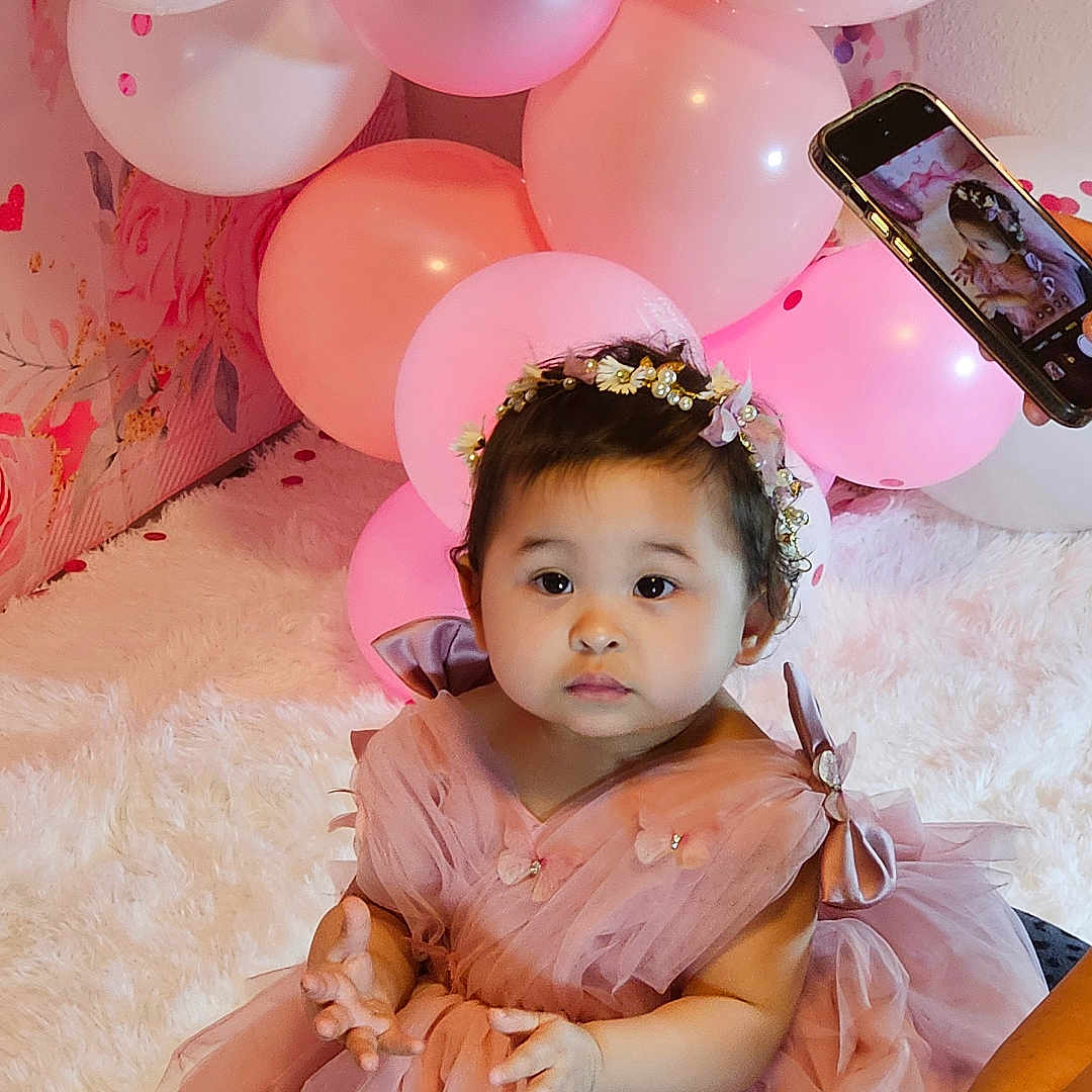 Zoey is registered to the contest to win money with this photo: adorable, baby, balloons, celebration, child, cute, decorations, floral, hands, headband, indoor, infant, party, photography, pink_dress, portrait, sitting, smartphone, tulle, white_rug