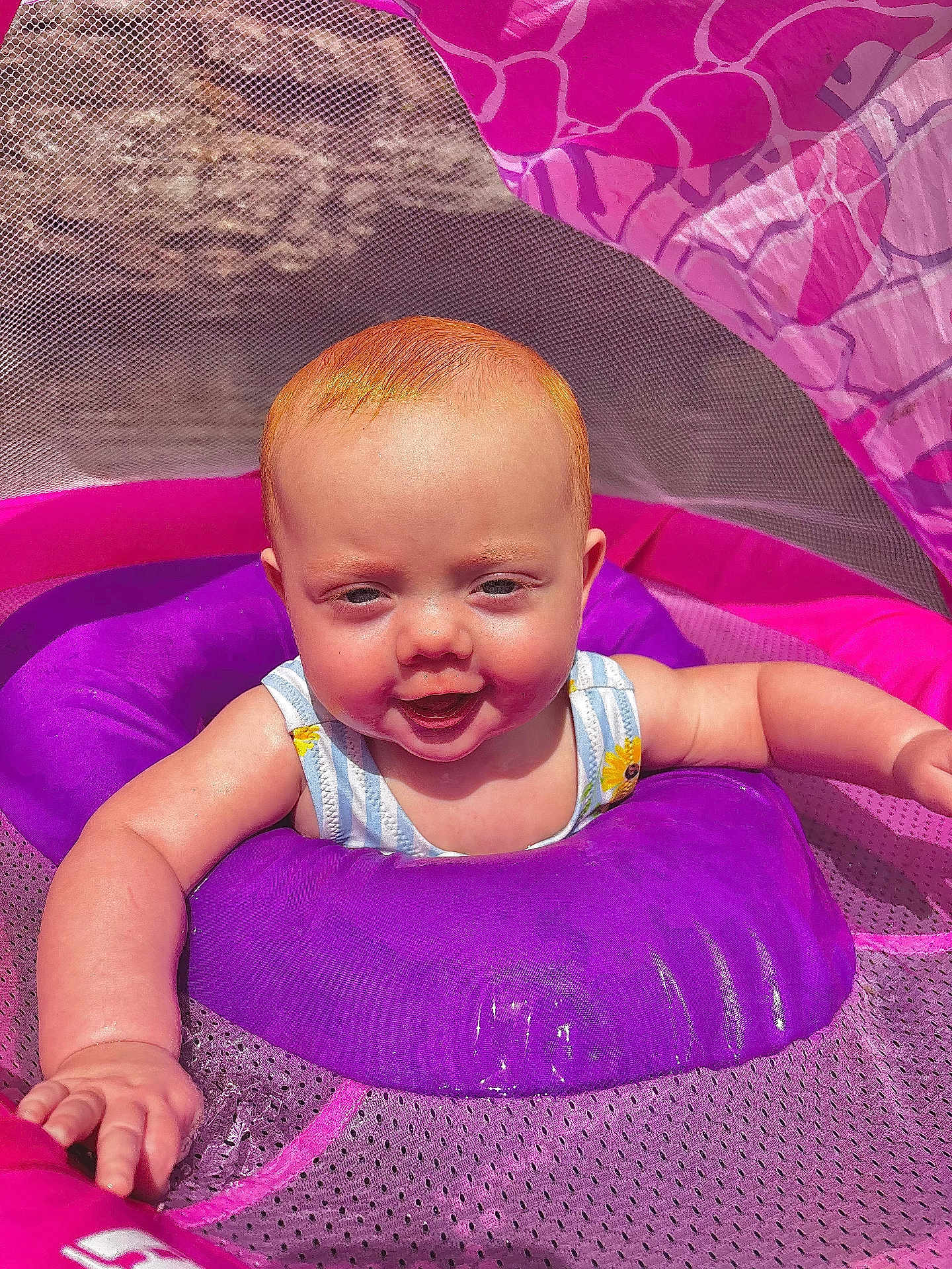 Lyra is registered to the contest to win money with this photo: baby, child, infant, red_hair, purple_inflatable, pink_mesh, sunlight, outdoor, smiling, happy, water_toy, summer, playtime, cute, young_child, baby_swim_ring, bright_colors, reaching_out, closeup, fun