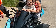 Alex joined the competition — help win amazing prizes! child, motorcycle, toddler, braids, outdoor, daytime, person, vehicle, blue_motorcycle, street, adult, casual_clothing, helmet_absent, playful, grip, handlebars, two_children, background, sunny, side_mirror