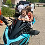 child, motorcycle, toddler, braids, outdoor, daytime, person, vehicle, blue_motorcycle, street, adult, casual_clothing, helmet_absent, playful, grip, handlebars, two_children, background, sunny, side_mirror