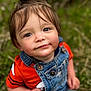 toddler, child, face, portrait, denim_overalls, orange_shirt, top_knot, grass, outdoor, nature, curious, cute, young_child, person, casual_clothing, sitting, daylight, skin, hair, eyes