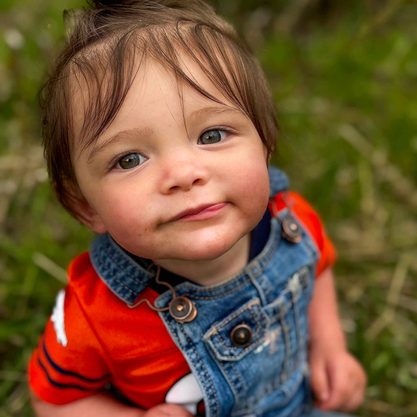 Xamiah is registered to the contest to win money with this photo: casual_clothing, child, curious, cute, daylight, denim_overalls, eyes, face, grass, hair, nature, orange_shirt, outdoor, person, portrait, sitting, skin, toddler, top_knot, young_child