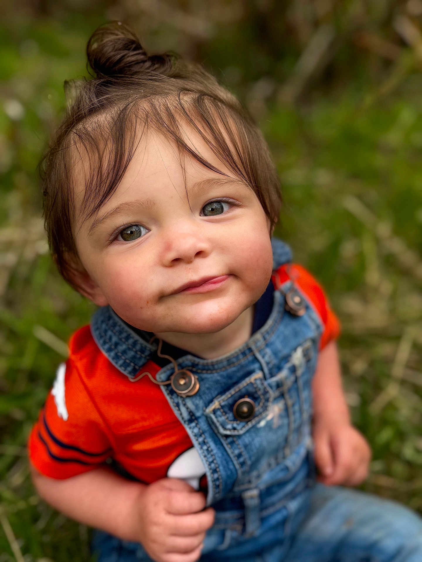 Xamiah is registered to the contest to win money with this photo: toddler, child, face, portrait, denim_overalls, orange_shirt, top_knot, grass, outdoor, nature, curious, cute, young_child, person, casual_clothing, sitting, daylight, skin, hair, eyes