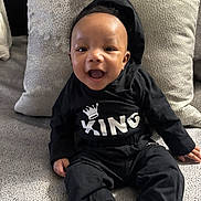 Jeremiah is registered to the contest to win money with this photo: baby, child, smiling, hoodie, black_clothing, sitting, pillow, couch, happy, indoor, infant, cute, portrait, person, comfortable, cozy, young_child, apparel, fabric, expression
