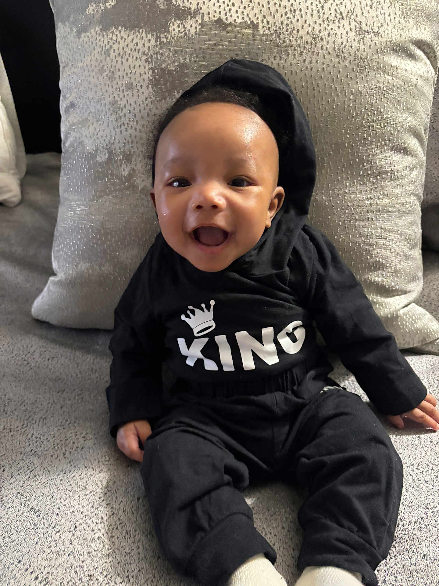 Jeremiah is registered to the contest to win money with this photo: baby, child, smiling, hoodie, black_clothing, sitting, pillow, couch, happy, indoor, infant, cute, portrait, person, comfortable, cozy, young_child, apparel, fabric, expression