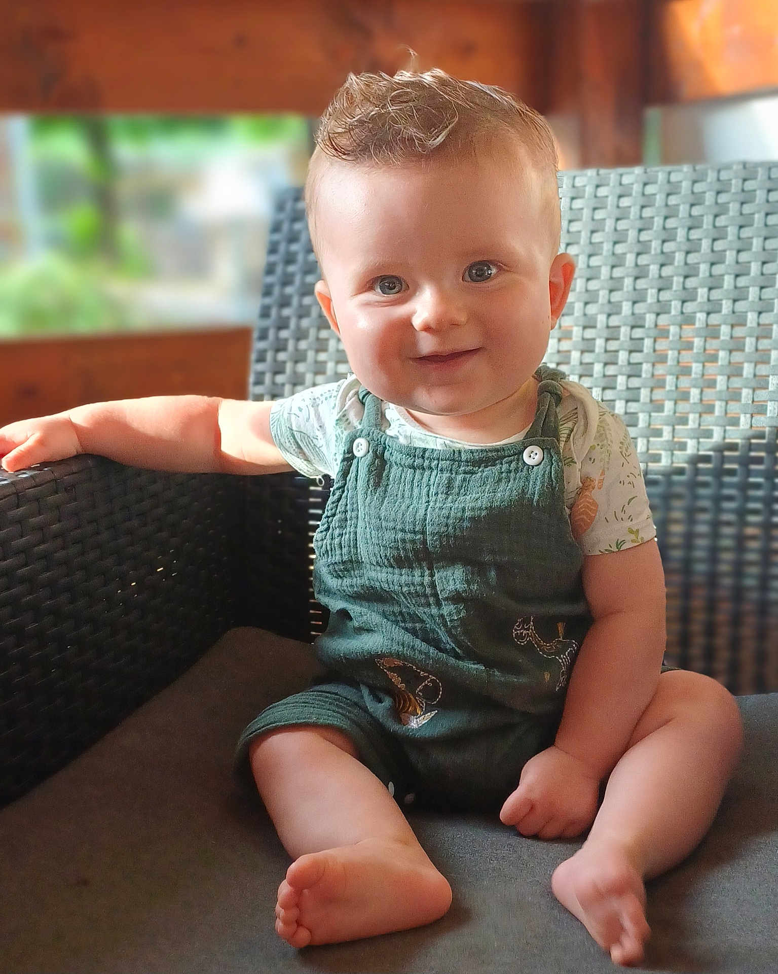 Côme participe au concours pour gagner de l'argent avec cette photo : baby, child, smiling, face, blue_eyes, green_overalls, short_hair, sitting, wicker_chair, armrest, bare_feet, indoor, natural_light, portrait, cute, happy, infant, casual_clothing, soft_background, wooden_wall